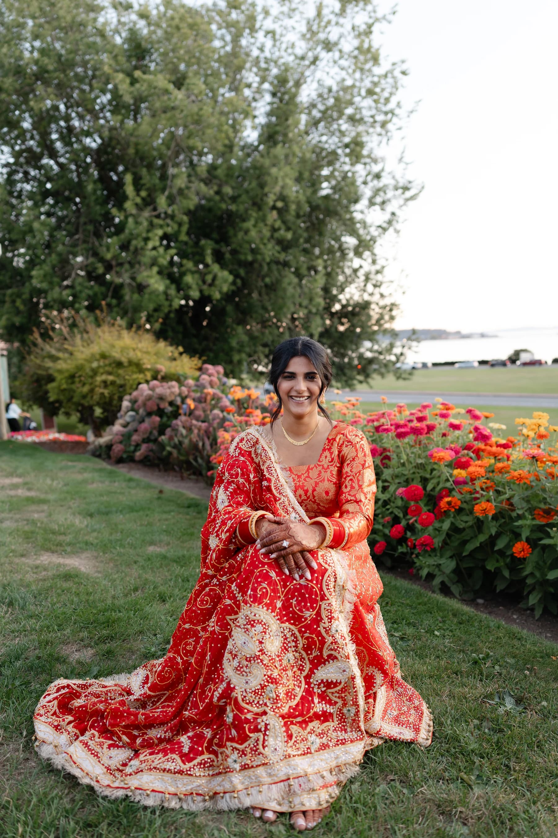 A woman in an embroidered red outfit seated beside red flowers in a garden.